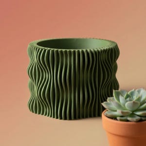 Puede incluir: Una maceta verde con bordes ondulados con una suculenta en una maceta de terracota. La maceta tiene forma cilíndrica con líneas verticales y onduladas. La suculenta es de color verde claro y está en una maceta pequeña y redonda de terracota.