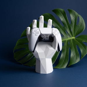 May include: A white, geometric, low-poly hand sculpture acting as a stand, holding a white and black wireless video game controller. The controller features a central touchpad, dual analog sticks, and standard directional buttons on its face.