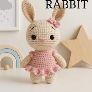 Peut inclure: Une poupée lapin au crochet avec une robe rose et un nœud rose. La poupée a des yeux noirs et un nez rose. Le texte "CUTE RABBIT" est au-dessus de la poupée. Une étoile en bois et un arc-en-ciel sont en arrière-plan.
