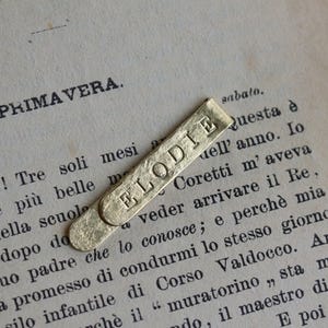Może przedstawiać: Prostokątna spinka do krawata w kolorze złotym z wytłoczonym imieniem ELODIE wielkimi literami. Metalowa powierzchnia ma matowe, teksturowane wykończenie z zaokrąglonymi końcami i jest umieszczona ukośnie na stronie z wydrukowanym tekstem.