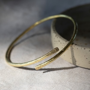 May include: A gold-toned bangle bracelet with a simple, minimalist design. The bracelet is made of a single, continuous piece of metal that is bent into a circle. The bracelet has a slightly hammered texture, giving it a rustic look.