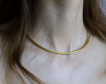 Puede incluir: Un collar tipo gargantilla de tono dorado que presenta un diseño de banda circular simple con un acabado de superficie texturizado y ligeramente irregular. El collar descansa cerca de la base del cuello, manteniendo un perfil fino, redondeado y uniforme.