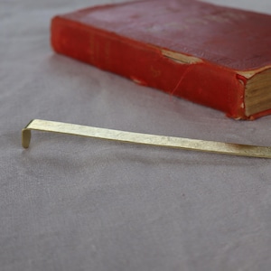 May include: A flat, elongated gold-colored metal bookmark with a simple hook-shaped end. The bookmark has a smooth, metallic finish and is positioned horizontally, showcasing its slender, rectangular profile and clean, minimalist design.