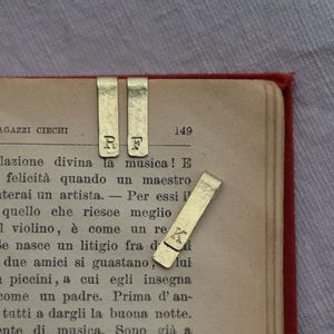 May include: Three rectangular gold-toned metal bookmarks with a brushed finish. Two bookmarks are positioned vertically with the stamped letters R and F, while one bookmark is placed diagonally featuring the stamped letter K on the surface.