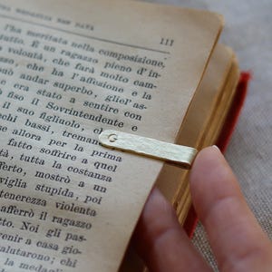 Op de afbeelding: Een rechthoekige bladwijzerclip van goudkleurig metaal met een getextureerd oppervlak en de letter G gestempeld op het bovenste uiteinde. De bladwijzer is bevestigd aan de pagina van een open boek met gedrukte Italiaanse tekst.