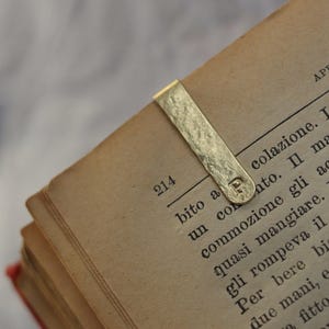 Könnte beinhalten: Ein goldfarbenes Lesezeichen aus Metall mit einer strukturierten, gehämmerten Oberfläche und einem abgerundeten Ende. Der Buchstabe P ist nahe dem unteren Ende eingeprägt, das an der oberen Ecke einer offenen Buchseite mit schwarzem Text klemmt.