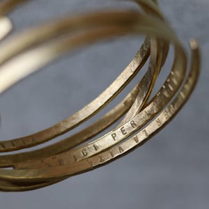 Op de afbeelding: Close-up van meerdere goudkleurige armbanden. Elke armband is gegraveerd met de woorden "PER LA VITA AMICIA". De armbanden zijn op elkaar gestapeld, waardoor een gelaagd effect ontstaat. Het metaal heeft een getextureerd, gehamerd uiterlijk.