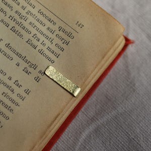 Op de afbeelding: Een rechthoekige bladwijzer van goudkleurig metaal met een getextureerd, gehamerd oppervlak. De bladwijzer is bevestigd aan de rand van een open boekpagina met gedrukte zwarte tekst en het paginanummer 147 in de bovenhoek.