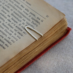 Pode incluir: Um marcador de página de metal prateado em forma de U colocado na página de um livro aberto com texto impresso em italiano. O marcador tem um acabamento fosco e um design simples e limpo, apoiado sobre a borda superior do papel.