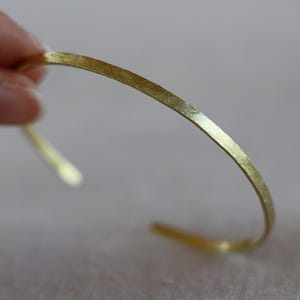 Peut inclure: Un bracelet manchette ouvert de couleur or. Le bracelet a une surface légèrement texturée et un design minimaliste. Les extrémités ouvertes permettent de le porter facilement. Ce bijou est simple et élégant.
