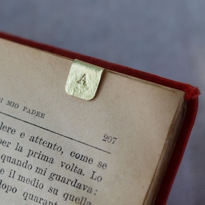 Op de afbeelding: Een goudkleurige metalen bladwijzer met een getextureerd oppervlak en de letter A in het midden gestempeld. De rechthoekige clip is bevestigd aan de bovenrand van een pagina in een open boek met vergeeld papier en zwarte gedrukte tekst.