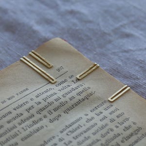 Può includere: Primo piano di diverse graffette dorate su una pagina di libro vintage. Il libro è aperto a pagina 207, con testo italiano visibile. Le graffette sono rettangolari con un design a doppia barra.