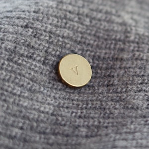 Może przedstawiać: Zbliżenie złotego, okrągłego guzika z literą "V" wygrawerowaną na powierzchni. Guzik umieszczony jest na tle szarej, teksturowanej dzianiny, tworząc kontrast kolorów i faktur.