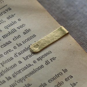 Könnte beinhalten: Ein goldfarbenes Lesezeichen aus Metall mit gehämmertem Finish und einem kleinen herzförmigen Ausschnitt am abgerundeten Ende, das am Rand einer offenen Buchseite befestigt ist, wobei schwarzer Text in Serifenschrift unter dem Metall sichtbar ist.