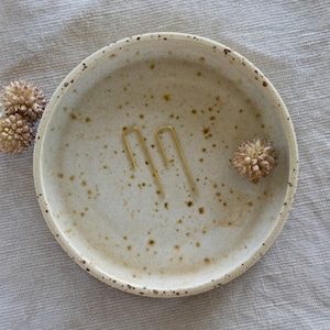 Può includere: Due orecchini minimalisti dorati a forma di U aperta su un piatto in ceramica bianco sporco con macchie.