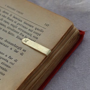 Può includere: Un segnalibro a clip in metallo dorato con la lettera M impressa sull'estremità arrotondata. La clip è fissata sul bordo di un libro aperto con pagine ingiallite e vissute, contenenti testo stampato in italiano, incluso il numero 147.