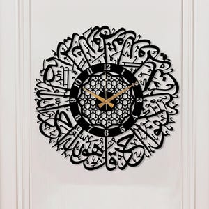 Könnte beinhalten: Schwarze, kreisförmige Wanduhr mit arabischer Kalligraphie. Die Uhr hat eine goldfarbene Mitte mit einem dekorativen Muster und schwarzen Ziffern. Sie ist an einer weißen Wand über einem Holztisch angebracht.