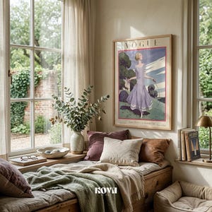 May include: A cozy window seat with a framed Vogue poster featuring a woman in a lavender dress. The scene includes decorative pillows, a green blanket, a vase with greenery, and a small lamp, creating a relaxing interior design.