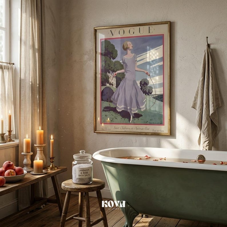 May include: A bathroom scene featuring a vintage Vogue poster in a gold frame, a green clawfoot bathtub, and a wooden stool with a jar of bath salts. Lit candles and a bowl of fruit add to the relaxing ambiance.