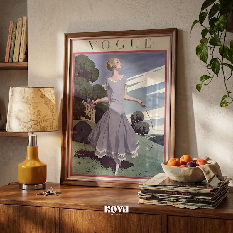 May include: A framed vintage Vogue magazine cover art print. The artwork features a woman in a flowing lavender dress, holding a rope, set against a landscape. The frame is a warm brown, and the print is displayed on a wooden cabinet.