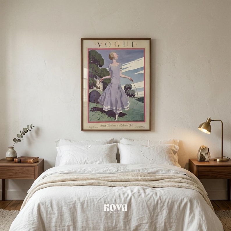 May include: A framed vintage "Vogue" magazine cover art print. The artwork features a woman in a lavender dress with a hoop, set against a green landscape and blue sky. The print is displayed above a bed with white linens and wooden nightstands.