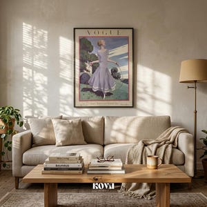 May include: A living room scene featuring a framed vintage Vogue magazine cover. The artwork depicts a woman in a lavender dress. A beige sofa with pillows, a wooden coffee table with books, and a floor lamp complete the room's decor.