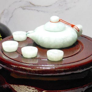 May include: A small, pale green jade tea set, including a teapot and four cups, sits on a dark wooden tray. The teapot has a red string tied to the lid. The set is displayed on a dark, glossy wooden stand.