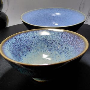 Puede incluir: Dos cuencos de cerámica con un esmalte azul moteado. Los cuencos tienen un borde marrón oscuro y un exterior azul oscuro. El interior de los cuencos es de color azul claro. Los cuencos están sobre una superficie negra.