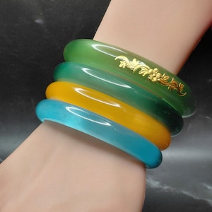 Pode incluir: Quatro pulseiras coloridas empilhadas em um pulso. As pulseiras são verdes, amarelas e azuis. A pulseira verde tem um design floral dourado. As pulseiras são feitas de um material translúcido.