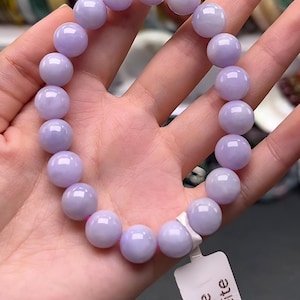 Könnte beinhalten: Ein elastisches Armband aus glatten, runden, hellvioletten Jadeit-Perlen. Die Perlen haben eine polierte, glänzende Oberfläche und sind auf einer flexiblen Schnur aufgereiht. Ein kleines weißes rechteckiges Etikett trägt den Text Purple Jadeite.