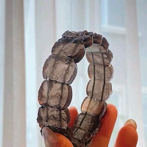 May include: A smoky quartz bracelet featuring large, rounded, faceted beads. The bracelet is a translucent gray color and appears to be strung together. The bracelet is held up against a soft, blurred background.