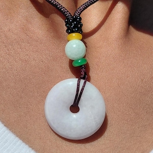 Könnte beinhalten: Eine weiße Jade-Anhänger-Halskette mit einem kreisförmigen, donut-förmigen Anhänger. Die Halskette hat eine dunkelbraune Kordel und ist mit farbigen Perlen in Gelb, Grün und Schwarz verziert. Der Anhänger ist hellcremeweiß.