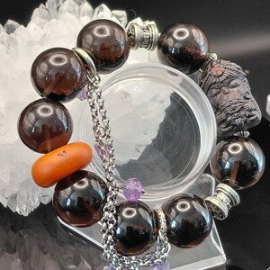 May include: A bracelet featuring dark brown translucent spherical beads, a single smooth orange oval bead, and a carved dark textured bead. The piece includes silver-toned metal spacers, a silver chain link charm, and small faceted purple beads.