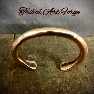 Op de afbeelding: Een gepolijste koperen manchetarmband met een open ontwerp. De armband heeft een gladde, afgeronde band en uitlopende uiteinden. De tekst "Tribal Art Forge" staat in rood script boven de armband.