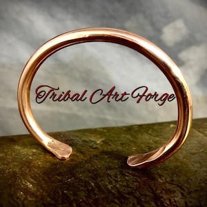 May include: A simple copper bracelet with a hammered finish. The bracelet is open and has a slightly curved shape. The text "Tribal Art Forge" is visible in the reflection of the bracelet.