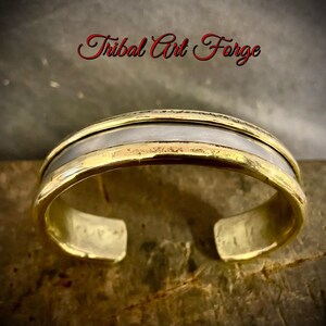 Può includere: Un bracciale rigido con un esterno in metallo dorato lucido e un inserto in metallo argentato liscio al centro. Il testo Tribal Art Forge è visualizzato in corsivo rosso sopra il bracciale.