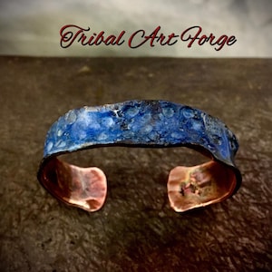 May include: A handcrafted copper cuff bracelet with a textured, blue enamel finish. The open-ended bracelet features a rustic, organic design, with the text "Tribal Art Forge" in red script above the bracelet.