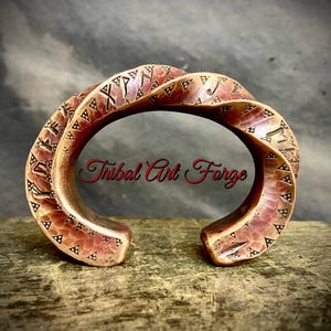 Peut inclure: Un bracelet manchette en métal cuivré avec une texture torsadée et martelée. La surface présente des caractères runiques gravés et de petits motifs triangulaires. Le texte Tribal Art Forge est superposé en rouge au centre du bracelet.