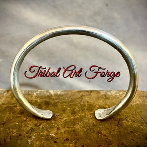 Pode incluir: Uma pulseira de metal cor de prata com uma banda lisa e arredondada e extremidades ligeiramente achatadas e alargadas. O texto "Tribal Art Forge" está sobreposto em letras vermelhas e pretas no centro da pulseira circular aberta.