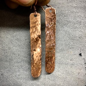 May include: A pair of copper-colored, hammered metal earrings. Each earring is a long, rectangular shape with a textured surface and a small hole at the top for the ear wire. The earrings are held against a gray background.