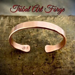 Peut inclure: Un bracelet manchette de couleur cuivre avec un design ouvert. Le bracelet a une surface lisse et légèrement incurvée et est estampillé d'un petit motif à une extrémité. Les mots "Tribal Art Forge" sont écrits en script rouge au-dessus du bracelet.