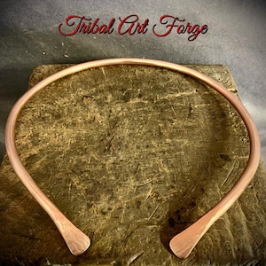 Puede incluir: Un collar gargantilla abierto de color cobre. El collar es una banda lisa y curvada con extremos aplanados. El texto "Tribal Art Forge" está en escritura roja encima del collar, sobre una superficie marrón texturizada.