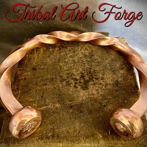 Könnte beinhalten: Ein kupferfarbenes, gedrehtes Metallarmband mit abgerundeten Enden. Es wird auf einer strukturierten, braunen Holzoberfläche präsentiert. Der Text "Tribal Art Forge" steht in roter Schrift darüber.