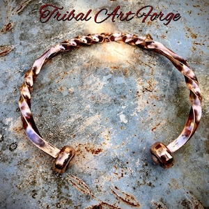 Könnte beinhalten: Ein kupferfarbenes Metallarmband mit gedrehtem Spiraldesign und abgerundeten, flachen Enden. Die Oberfläche des Metalls hat ein poliertes, reflektierendes Finish. Der Text "Tribal Art Forge" ist oben im Bild deutlich zu erkennen.