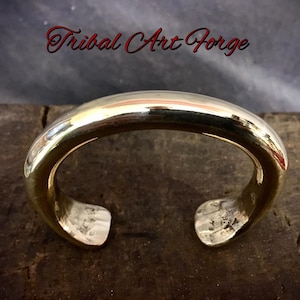 Peut inclure: Un bracelet manchette ouvert et poli, de couleur dorée, avec un design arrondi. Le bracelet est posé sur une surface sombre et texturée. Le texte "Tribal Art Forge" est affiché en script rouge au-dessus du bracelet.