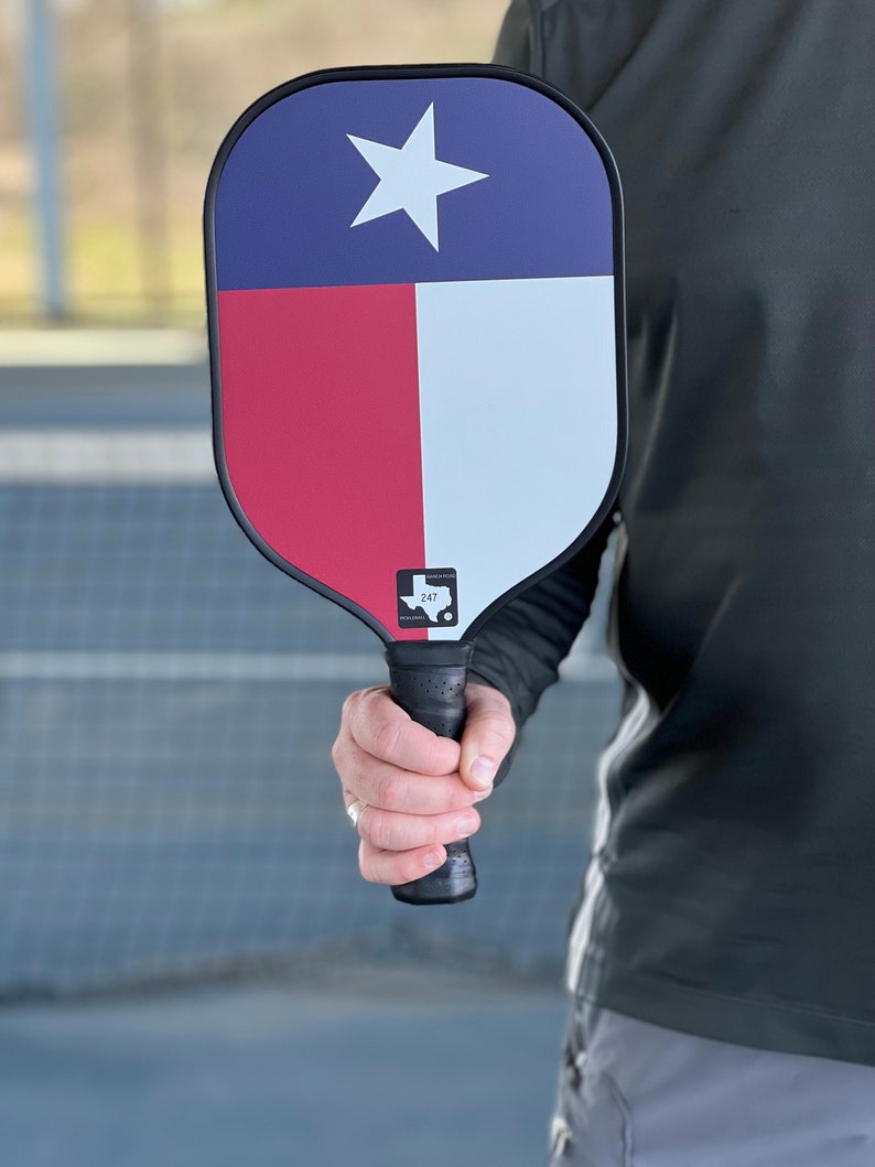 K&ouml;nnte beinhalten: Ein schwarzer und roter Pickleball-Schl&auml;ger mit einem Texas-Flaggen-Design. Der Schl&auml;ger hat einen schwarzen Griff und eine wei&szlig;e Umrandung des Bundesstaates Texas mit der Nummer 247 im Inneren.