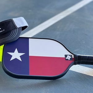 K&ouml;nnte beinhalten: Ein schwarz-wei&szlig;es Pickleball-Schl&auml;ger mit einem Texas-Flaggen-Design. Der Schl&auml;ger hat einen schwarzen Griff und einen wei&szlig;en Aufkleber mit einer Texas-Umrandung und dem Text "TX" darauf. Eine schwarz-wei&szlig;e Baseballkappe mit einem wei&szlig;en Netzr&uuml;cken und dem Text "Come and Dink It" liegt auf dem Schl&auml;ger. Ein gelber Pickleball befindet sich im Hintergrund.