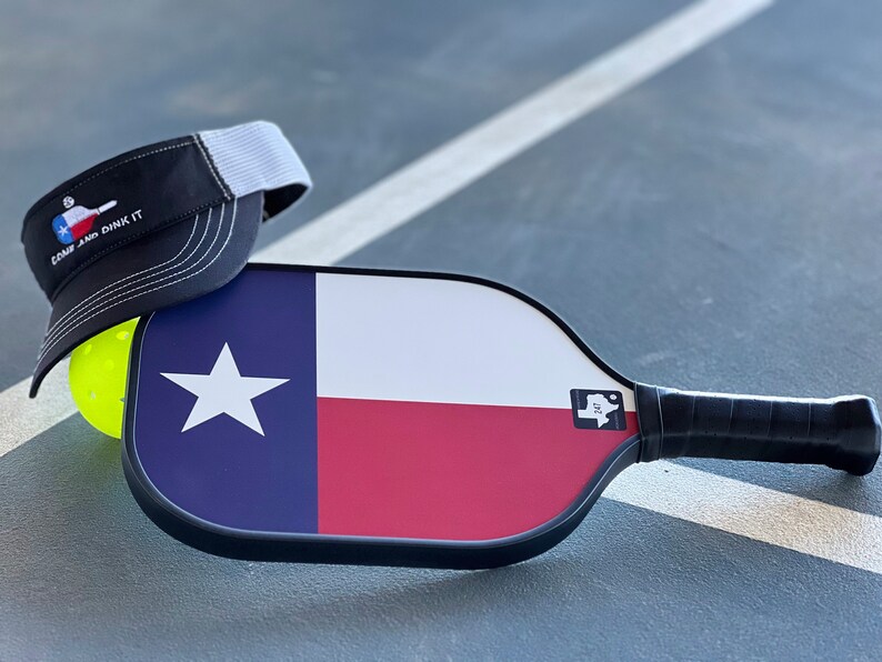 K&ouml;nnte beinhalten: Ein schwarz-wei&szlig;es Pickleball-Schl&auml;ger mit einem Texas-Flaggen-Design. Der Schl&auml;ger hat einen schwarzen Griff und einen wei&szlig;en Aufkleber mit einer Texas-Umrandung und dem Text "TX" darauf. Eine schwarz-wei&szlig;e Baseballkappe mit einem wei&szlig;en Netzr&uuml;cken und dem Text "Come and Dink It" liegt auf dem Schl&auml;ger. Ein gelber Pickleball befindet sich im Hintergrund.