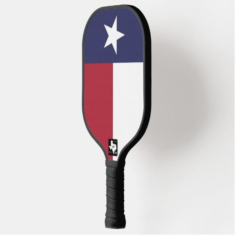 K&ouml;nnte beinhalten: Ein schwarzer und roter Pickleball-Schl&auml;ger mit einem Texas-Flaggen-Design. Der Schl&auml;ger hat einen schwarzen Griff und ein schwarz-wei&szlig;es Logo mit dem Bundesstaat Texas.