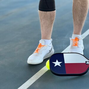 K&ouml;nnte beinhalten: Eine Person in wei&szlig;en Sneakers mit orangenen Schn&uuml;rsenkeln steht auf einem Pickleball-Platz mit einem Pickleball-Schl&auml;ger, der das Design der Flagge des Bundesstaates Texas tr&auml;gt. Ein gelber Pickleball liegt auf dem Platz.